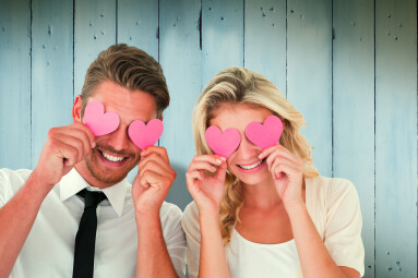 Attractive,Young,Couple,Holding,Pink,Hearts,Over,Eyes,Against,Wooden