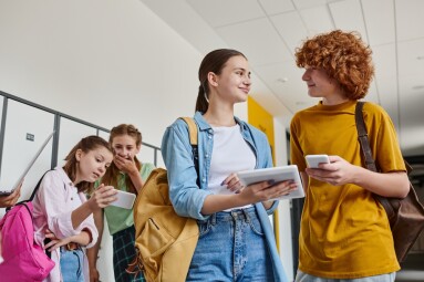 Happy,Teenage,Boy,And,Girl,Holding,Gadgets,Near,Classmates,In