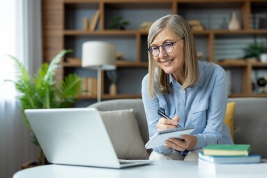 Mature,Woman,Studying,Online,With,Laptop,,Writing,Notes,,Smiling,While