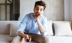 Bored,Young,Man,Holding,Tv,Remote,Control,While,Sitting,On