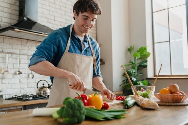 Young,Man,In,Apron,Standing,In,Home,Kitchen,Slicing,Vegetables
